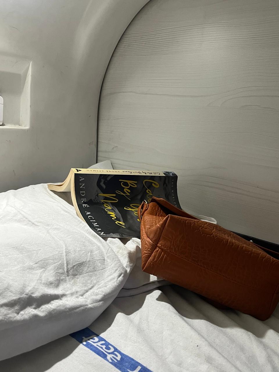 A book laying open with its face down on a white pillow on a white sheet. An orange bag laying flat next to the book is covering parts of its front cover. Visible writing reads 'Ca', 'By', 'Name', and the author's name, 'André Aciman'.