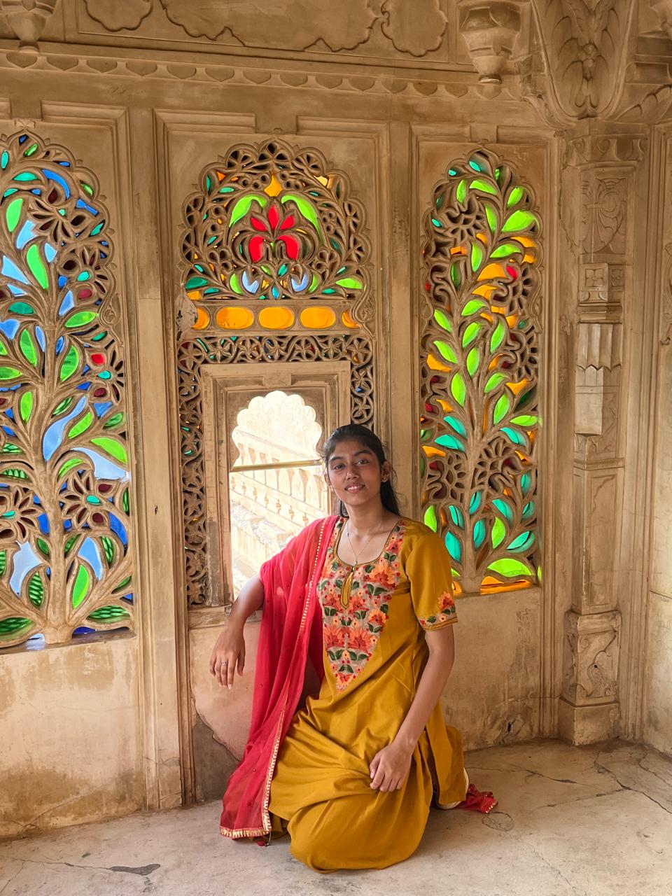 Picture of a girl in a yellow-ochre coloured kurta and salwar (type of pants) sitting on the floor facing the camera with a hand propped up on a small low window on a wall with floral stained glass designs.
